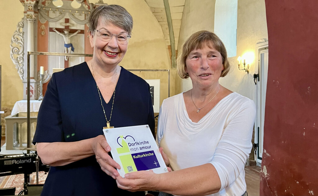 Landesbischöfin KristinaKühnbaum-Schmidt überreicht ein Schild "Dorfkirche mon amour"